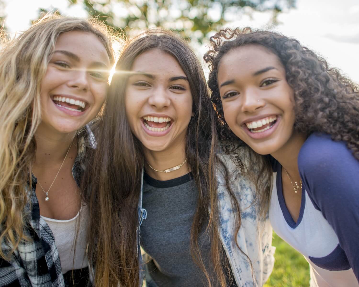 Three teenage girls outside on a sunny day. They are all smiling at the camera together while embracing.